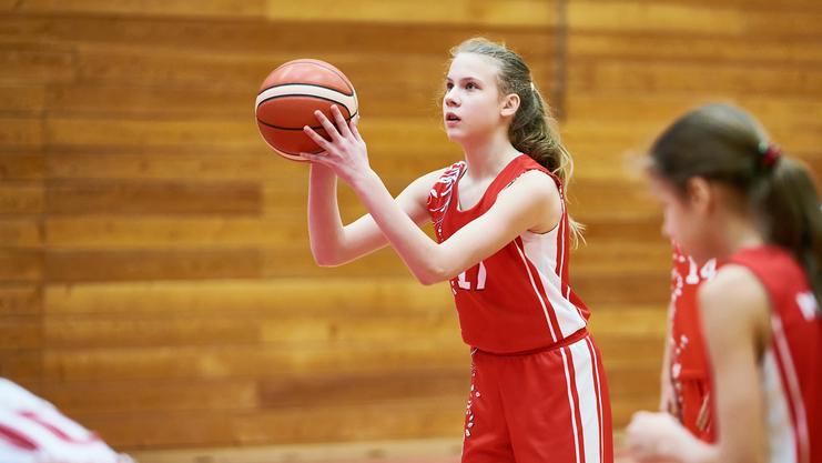 Mädchen in rotem Basketballtrikot bereitet sich auf einen Freiwurf mit Ball in der Hand an einer Holzwand.