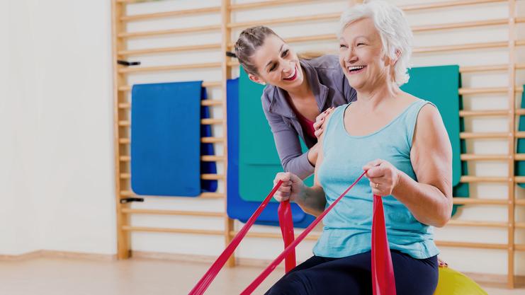 Frau mit grauen Haaren sitzt auf einem großen Gymnastikball, während eine Trainerin ihr mit engen Bändern hilft.
