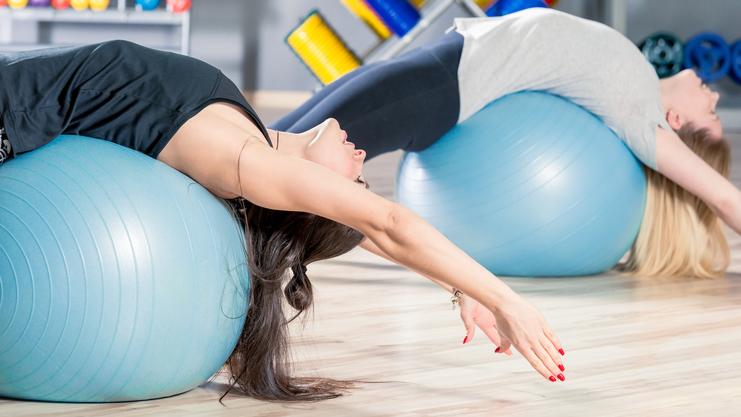 Zwei Frauen dehnen sich auf blauen Gymnastikbällen in einem Fitnessstudio mit Holzfußboden.