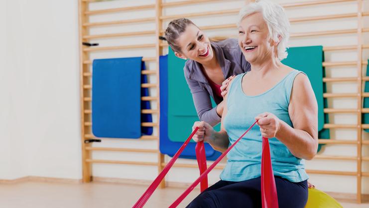 Frau mit grauen Haaren sitzt auf einem großen Gymnastikball, während eine Trainerin ihr mit engen Bändern hilft.