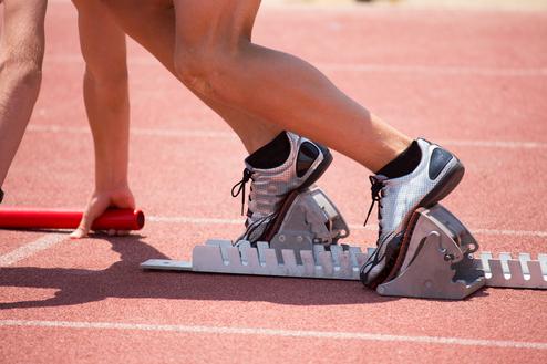Startposition eines Sprintläufers mit Fuß auf einem Startblock auf einer roten Laufbahn, Hand hält einen Staffelstab.