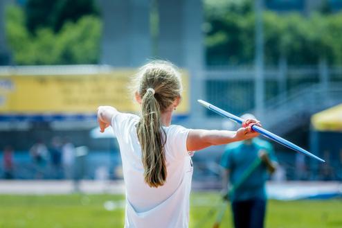 Mädchen wirft eine Sportspitze, trägt ein weißes T-Shirt und steht auf einer grünen Sportfläche.