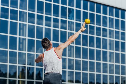 Sportler wirft einen Ball in die Luft, vor einer großen Glasfassade an einem sonnigen Tag.
