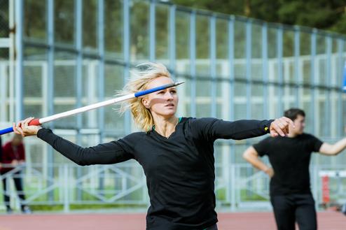 Athletin wirft im Sportoutfit einen Speer auf einer Laufbahn, im Hintergrund ist ein weiterer Sportler zu sehen.