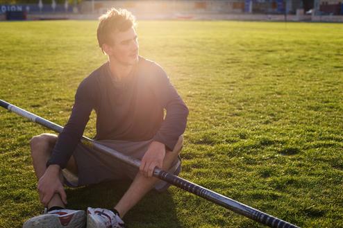 Junger männlicher Sportler sitzt auf einer Rasenfläche und hält einen Stab im Schatten des Sonnenlichts.