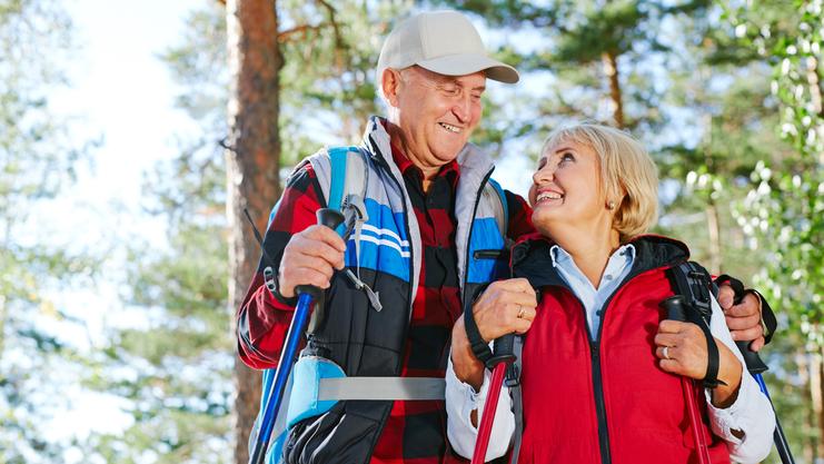 Ein älteres Paar steht in einem Wald, lächelt sich an und hält Wanderstöcke in den Händen.