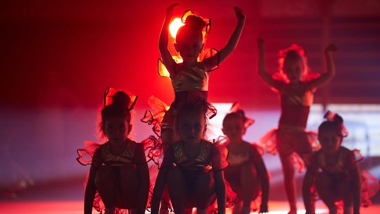 Sechs Kinder in goldenen Kostümen mit roten Rüschen posieren in rotem Licht auf der Bühne.